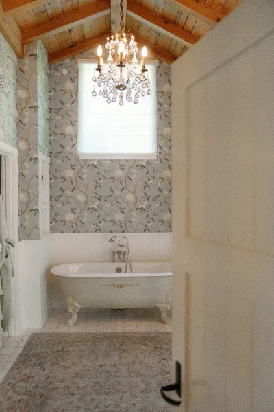 Getting ready bathroom with a crystal chandelier above a clawfoot bathtub, blue floral wallpaper, and wood vaulted ceiling over white wainscoting