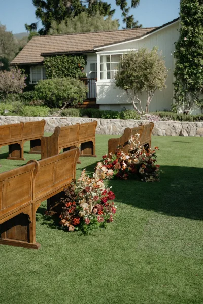 Ceremony aisle decor with outdoor ceremony seating, wooden pews lined by meadow florals and greenery on grass beside an ivy cottage stone wall.