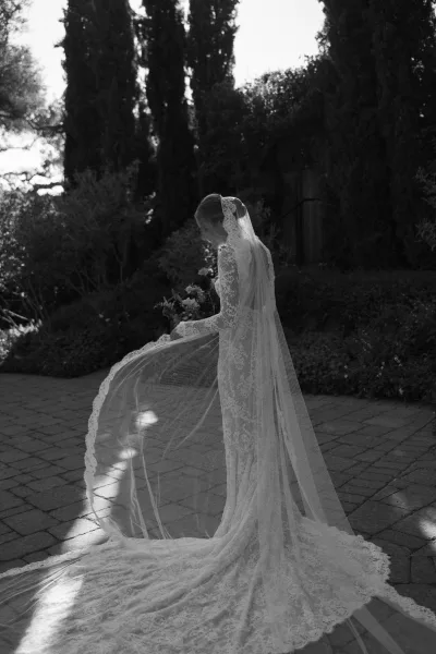 Bridal portrait of a bride in a lace wedding dress with long veil, looking down holding a bouquet on a sunlit stone patio garden