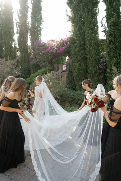 Bride with bridesmaids in a bridal veil shot, black dresses holding her long lace veil as she walks along a garden stone walkway