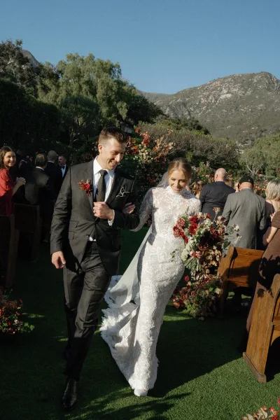 Wedding recessional as bride and groom walk the aisle, bouquet in hand, under a floral arch with guests, mountains, and blue sky behind