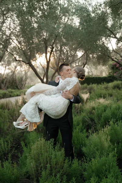 Wedding kiss portrait of groom carrying bride in a lace gown, her veil flowing as they dip-kiss in sunset-lit garden greenery