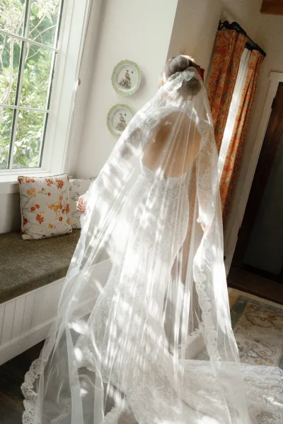 Bridal portrait of a bride in veil, showcasing a long lace veil train and backless gown by window light on a window seat with patterned curtains