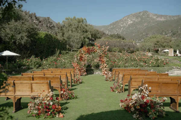 Ceremony setup with outdoor wedding ceremony floral arch, wooden pews and aisle florals on a lawn with mountain view and rose garden backdrop