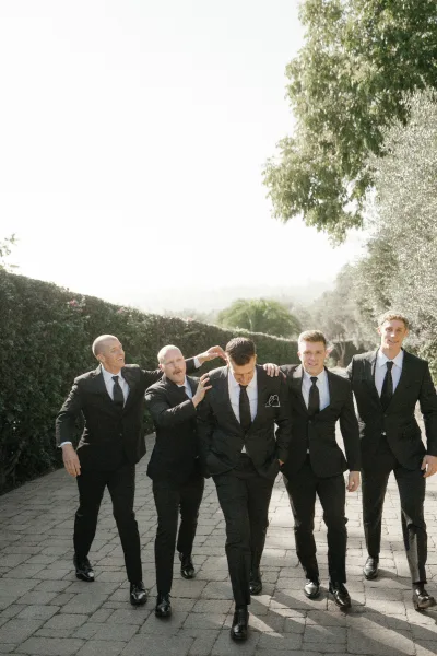 Groomsmen portrait of men in matching black suits and black ties walking and laughing on a stone paver path by tall hedges under bright sky