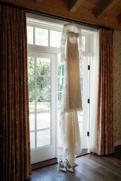 Wedding dress hanging by French doors in window light, lace wedding dress with long sleeves and white pointed-toe heels on the wood floor