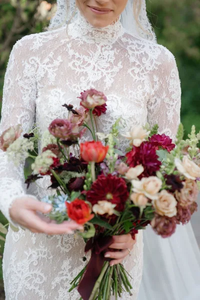 Bridal portrait of a bride in a high neck lace wedding dress, holding a burgundy bouquet with silk ribbon, framed by garden greenery
