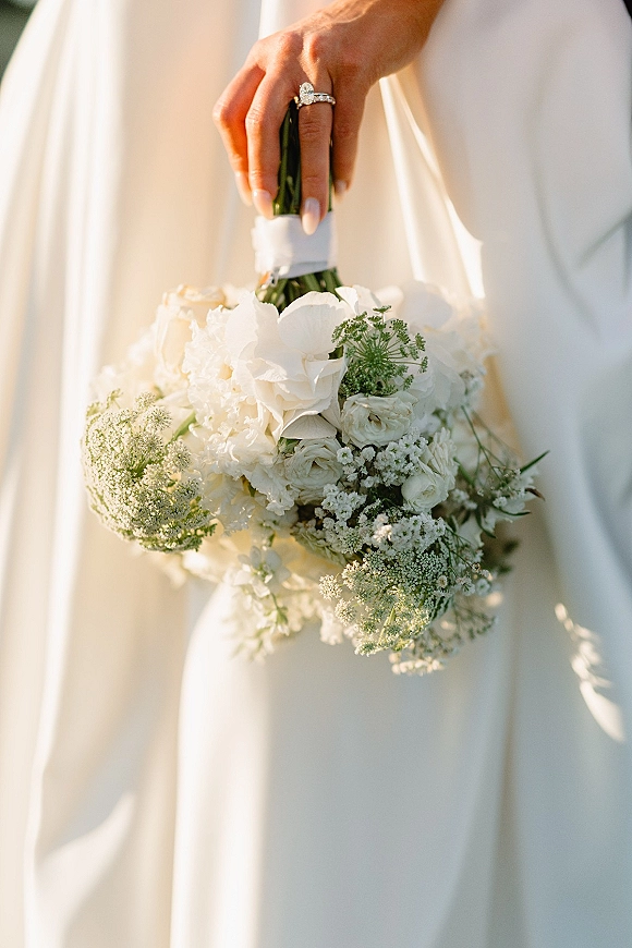 Bridal bouquet of white wedding bouquet blooms and greenery held in hand, ribbon-wrapped, with oval diamond ring and manicure in soft light