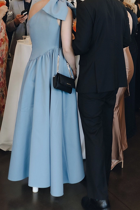 Wedding guest outfit in a blue wedding guest dress with bow shoulder, holding a cocktail glass beside a high-top table in a reception crowd