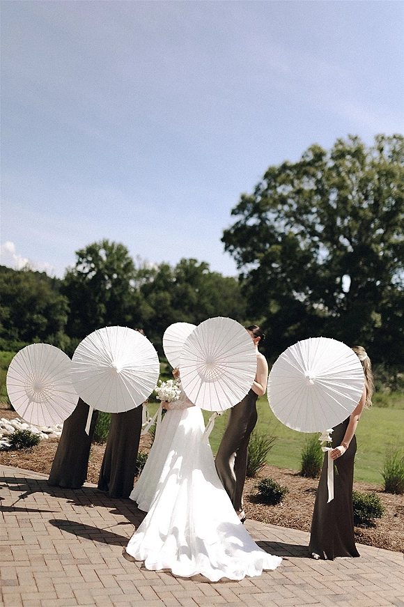 Bridal party portrait with bridesmaids with parasols, bride holding a bouquet as everyone hides faces on a sunny brick patio lawn