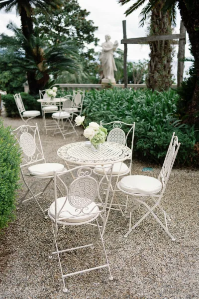 Reception lounge seating with a white wrought iron bistro table, chairs, and rose centerpiece in a lush garden courtyard with palms and statue