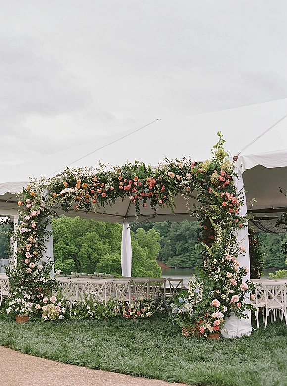Wedding ceremony arch with floral wedding arch roses, hydrangeas, and greenery garland under a white tent by a lakeside lawn