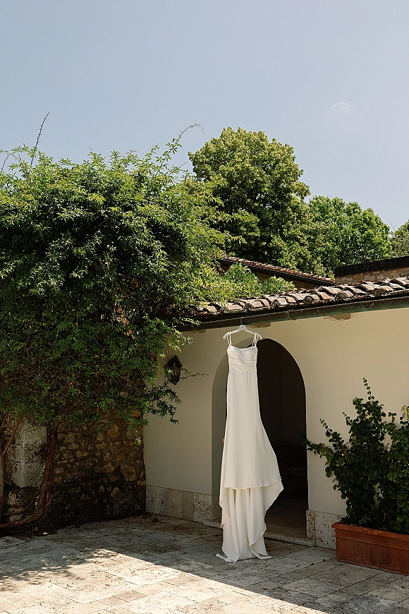 Wedding dress hanging from a doorway, simple wedding dress in a stucco courtyard with lantern, vines, terracotta planter, and blue sky