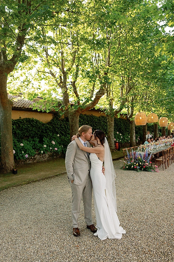 Wedding kiss as the bride and groom embrace beside a long banquet table with hanging lantern accents in a gravel courtyard garden