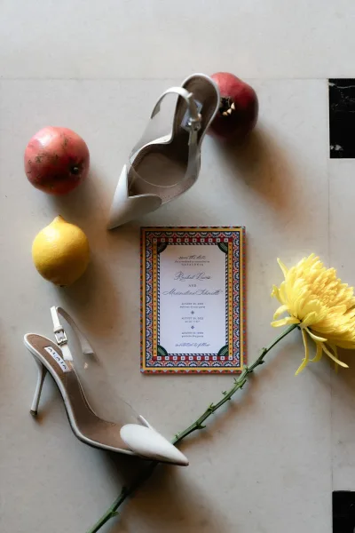 Wedding invitation flatlay with a save the date card, white slingback heels, pomegranates, lemon, and yellow chrysanthemum on marble surface