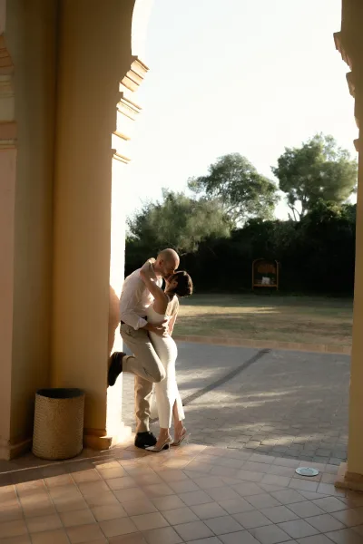 Wedding kiss portrait of bride and groom kissing in a dip, her white heels lifted, backlit beneath a stucco archway in a courtyard