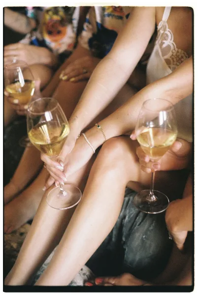 Bridesmaids toast with getting ready champagne, clinking white wine glasses over a couch and patterned rug in a cozy indoor room