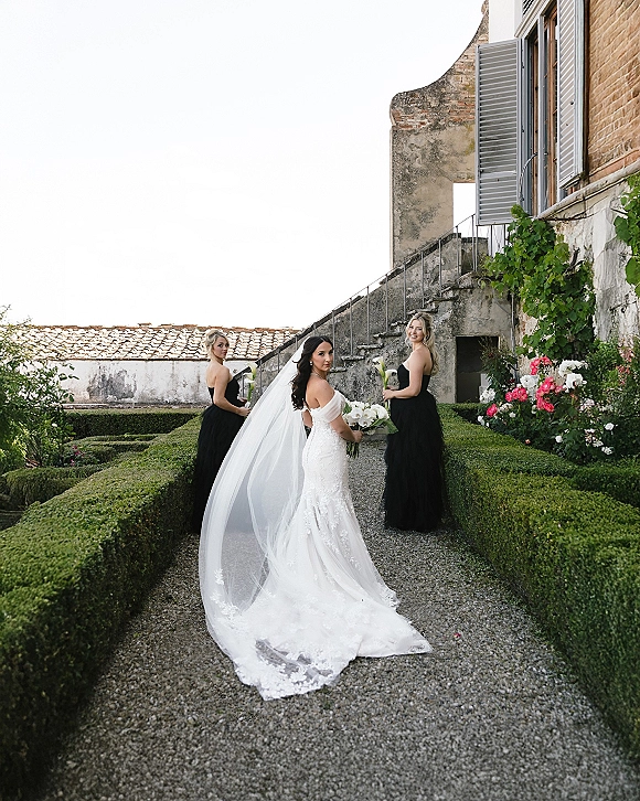 Bride with bridesmaids in black dresses holding white calla lily bouquets, bride in long veil and lace train on gravel path by ivy villa stairs