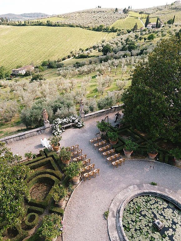 Outdoor ceremony setup with wood chairs and a white floral arch, set in a gravel courtyard beside a lily pond and vineyard hills