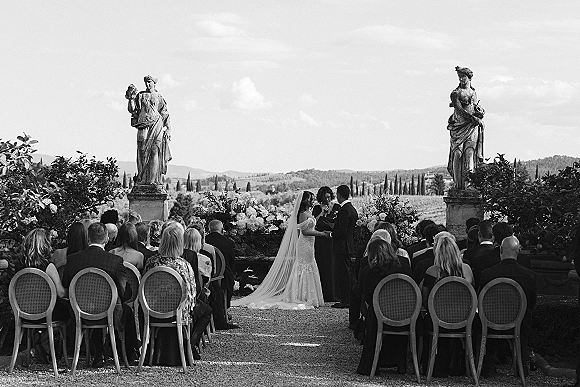 Wedding ceremony with bride in veil and groom in suit at an outdoor wedding ceremony on a garden terrace, floral-lined aisle, stone statues, hills beyond