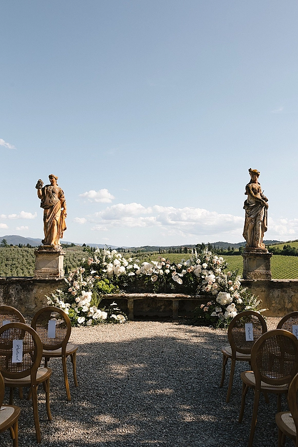 Ceremony setup for outdoor wedding ceremony with a white and blush floral arch, cane chairs, and reserved signs on a stone terrace overlooking vineyards