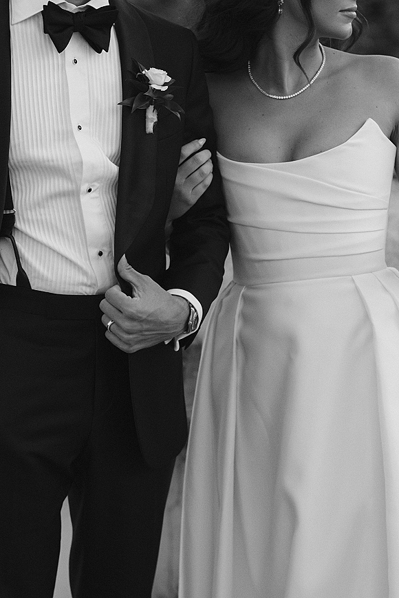 Wedding couple portrait of bride and groom arm in arm, her pearl necklace and strapless dress beside his tuxedo boutonniere on an outdoor walkway