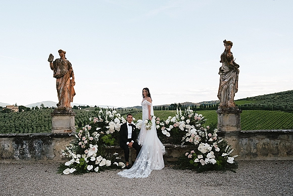Couple portrait of bride in an off-the-shoulder dress and veil beside groom in tuxedo on a stone terrace with vineyard hills backdrop