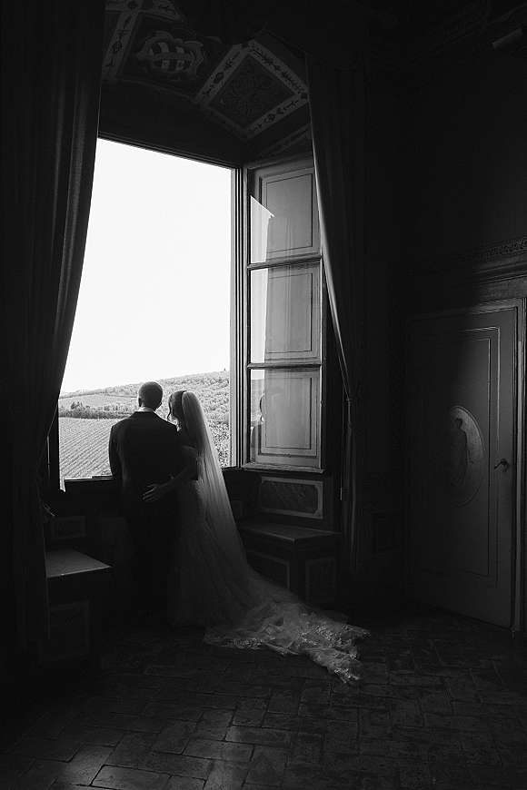 Couple portrait in black and white wedding portrait style, bride and groom silhouetted at a large window with veil and long train trailing.