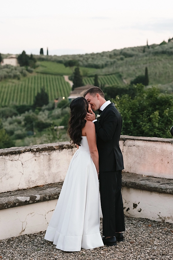 Wedding kiss portrait of bride and groom kiss on a stone terrace wall, groom in tuxedo with boutonniere, vineyards and hills behind