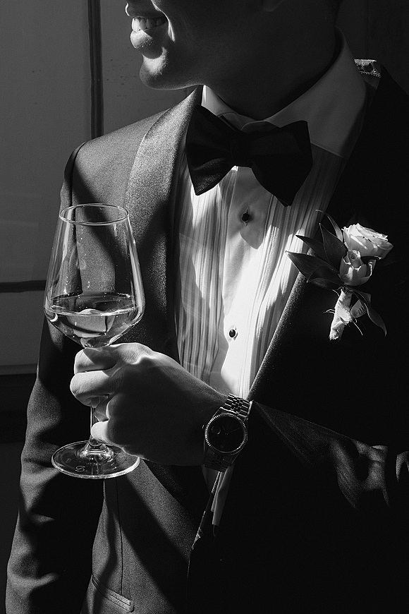 Groom portrait in a black tuxedo holding a wine glass, wearing a black bow tie, wristwatch, and rose boutonniere in window light