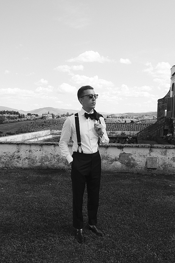 Groom portrait in suspenders with black bow tie and sunglasses, hands in pockets on a stone terrace overlooking rolling hills under clouds