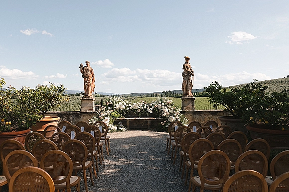 Ceremony setup with outdoor ceremony seating on a gravel aisle, rattan chairs and white-blush flowers on a stone terrace with vineyard hills beyond