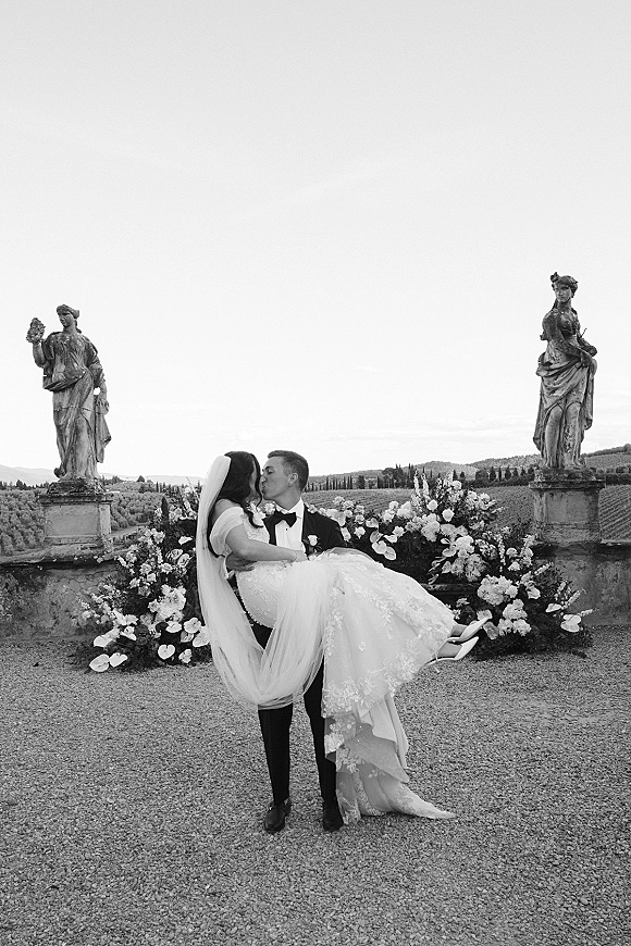 Wedding kiss portrait as the groom carrying bride in a lifted pose, her long veil flowing on a terrace with vineyard hills and stone statues