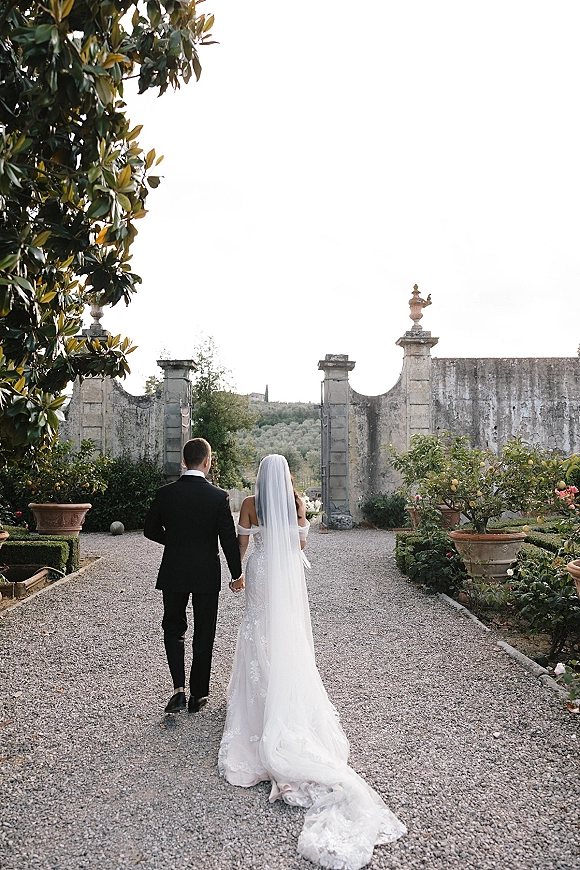 Couple portrait of newlyweds walking away hand in hand, bride in wedding dress with long veil and bouquet on a gravel garden path