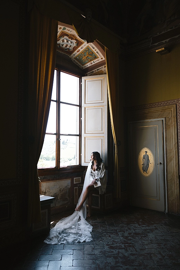 Bridal portrait of a bride by window, sitting on a windowsill in a lace train and veil, holding a stationery card in a historic room