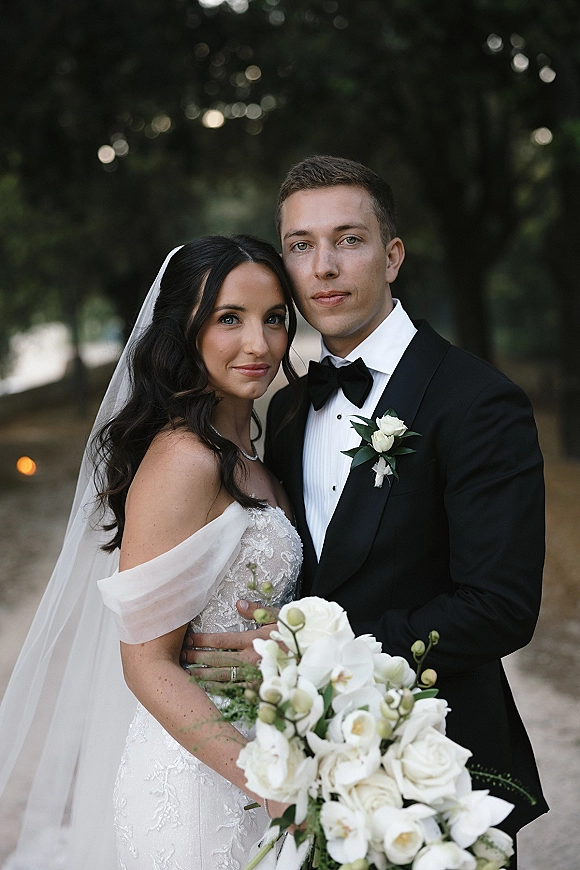 Couple portrait of bride and groom close up, embracing on a tree-lined path, bride in veil and holding a white bouquet beside groom in tuxedo