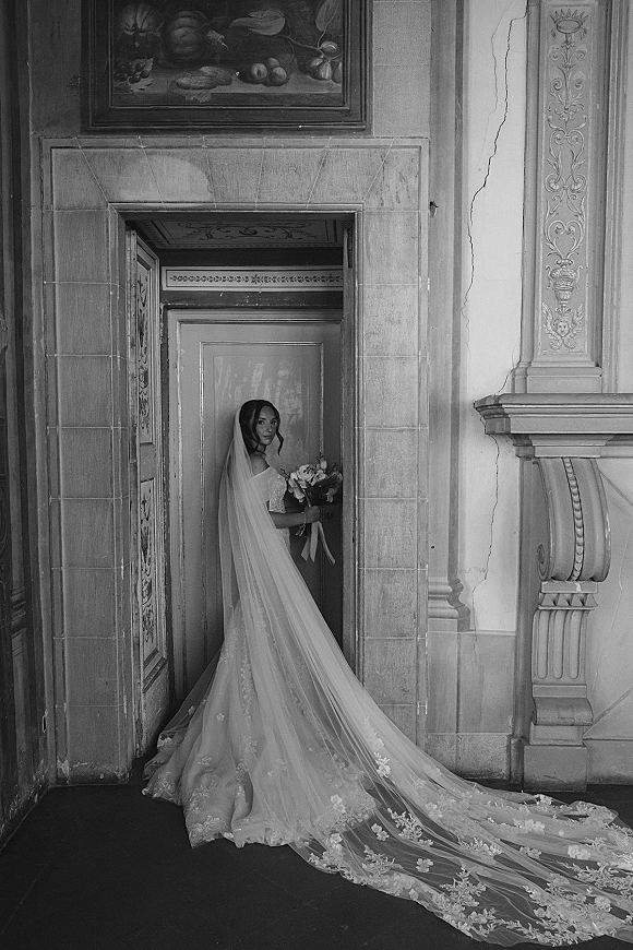 Bridal portrait in black and white of a bride holding a bouquet, long veil and lace dress trailing in an ornate interior doorway