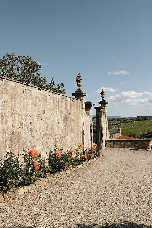 Outdoor wedding venue with stone wall wedding backdrop, gravel path to a wrought iron gate, rose bushes and urns under vineyard hills sky