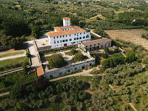 Wedding venue aerial view of a villa with terracotta roof tiles, stone courtyard walls and fountain, set amid olive groves and rolling hills