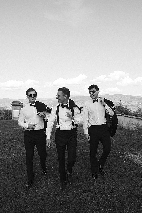 Groomsmen portrait of three men walking in sunglasses, tuxedo jackets and bow ties on a lawn by a stone wall with mountains behind