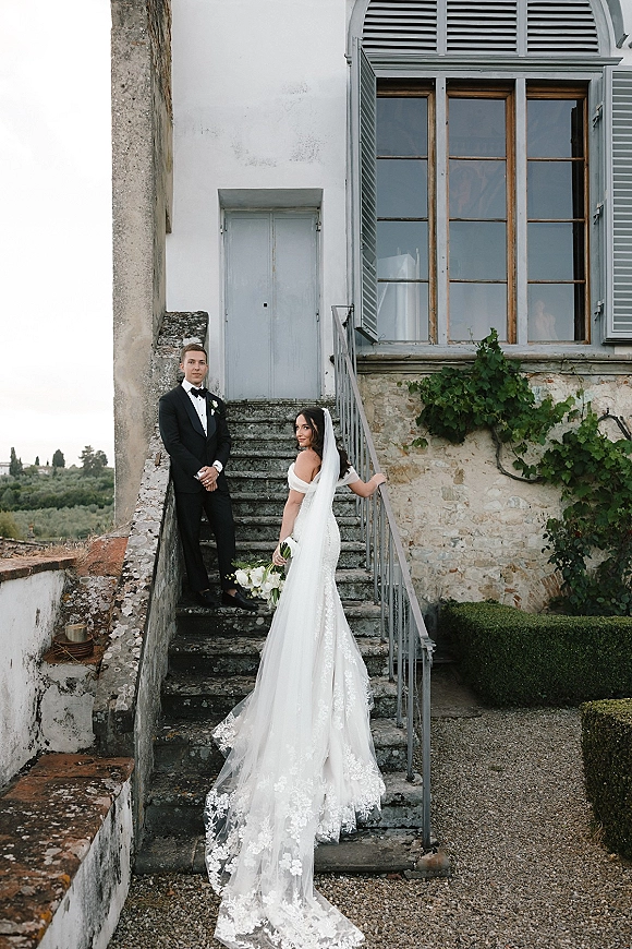 Couple portrait of bride and groom on stairs, her long veil and lace train draping down stone steps, holding a white bouquet by ivy wall