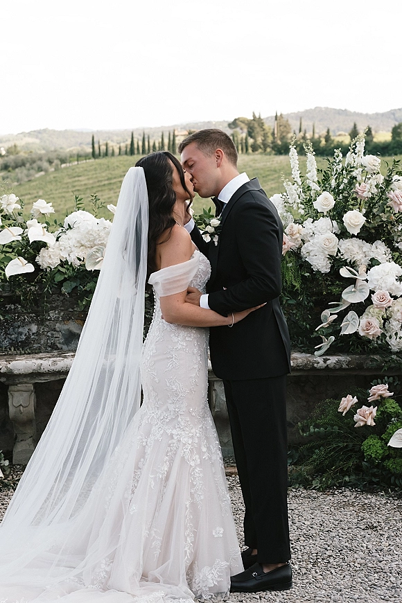 Wedding kiss portrait of bride and groom kissing, her long veil trailing as they embrace on a stone terrace with cypress trees and hills