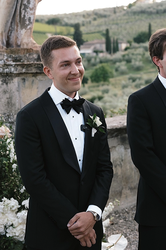 Groom portrait in a black tuxedo groom look with a white rose boutonniere, bow tie and lapel mic, standing by a stone wall garden terrace