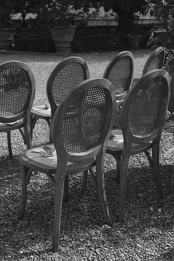 Ceremony seating with outdoor ceremony chairs featuring cane backs, paper programs and ribbon bows on gravel in a garden setting