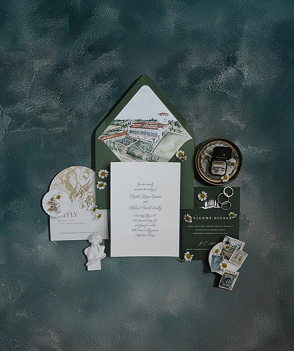 Wedding invitation suite with a letterpress wedding invitation, dark green envelope and wax seal, rings, pearls, and daisies on gray backdrop