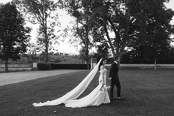 Wedding couple portrait in a black and white wedding portrait, bride holding bouquet and looking back as they walk on an estate lawn with trees