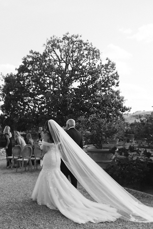 Wedding processional with bride walking down aisle in lace off-the-shoulder gown and long veil, holding bouquet on garden gravel path with guests seated
