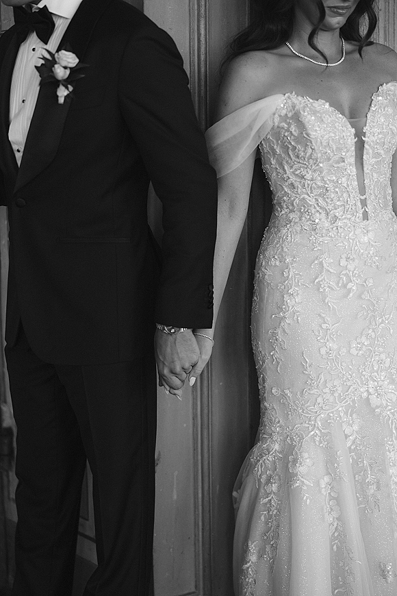 Couple portrait of bride and groom holding hands, showing off-shoulder lace wedding dress and tuxedo with boutonniere by wood door
