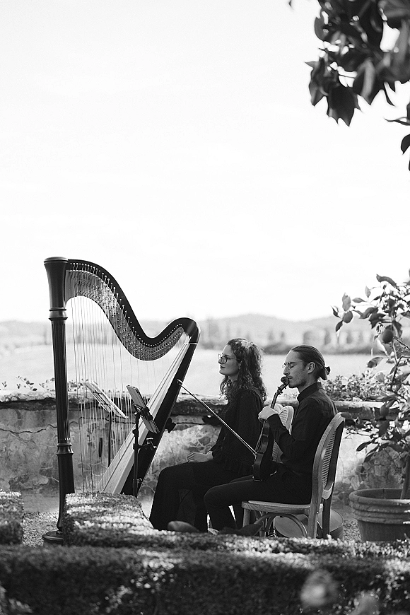 Wedding ceremony musicians, a harp and violin duo seated with sheet music by a stone garden wall overlooking a lake and hills