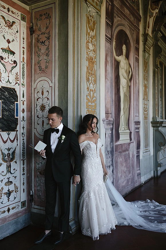 Wedding first look as bride and groom stand back to back holding hands, groom reading vow letter in ornate mural-lined hallway
